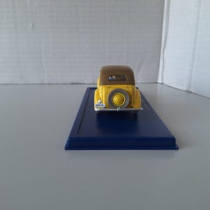 May include: A yellow and brown vintage car model with a spare tire on the back. The car is on a blue rectangular base. The model has a license plate. The car is a miniature collectible.