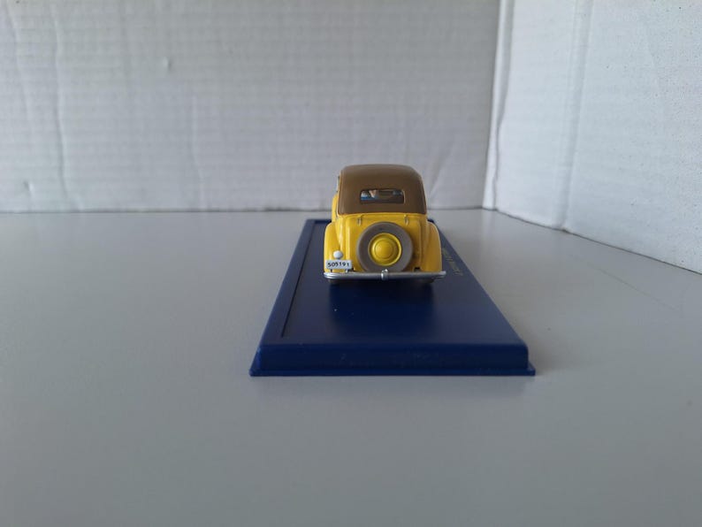 May include: A yellow and brown vintage car model with a spare tire on the back. The car is on a blue rectangular base. The model has a license plate. The car is a miniature collectible.