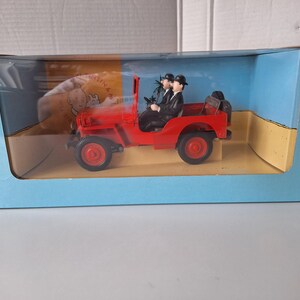May include: A red toy jeep with two figures in black suits and bowler hats. The jeep is in a blue box with a clear plastic front. The box has a cartoon image on the side.