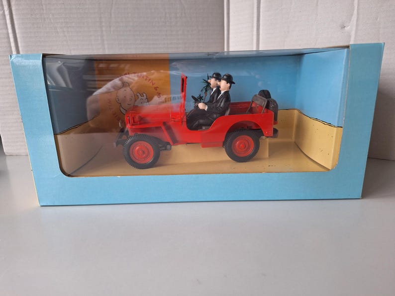 May include: A red toy jeep with two figures in black suits and bowler hats. The jeep is in a blue box with a clear plastic front. The box has a cartoon image on the side.