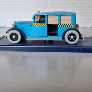 Może przedstawiać: Jasnoniebieski model zabytkowej taks&oacute;wki z czarno-ż&oacute;łtymi szachownicami. Zabawkowe auto ma figurkę kierowcy i stoi na ciemnoniebieskiej podstawie. Koła są szare z czarnymi środkami.