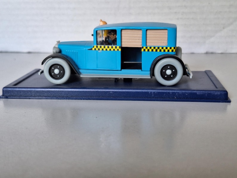 Może przedstawiać: Jasnoniebieski model zabytkowej taks&oacute;wki z czarno-ż&oacute;łtymi szachownicami. Zabawkowe auto ma figurkę kierowcy i stoi na ciemnoniebieskiej podstawie. Koła są szare z czarnymi środkami.