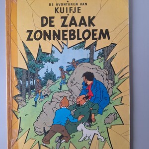 May include: A vintage comic book titled "De Zaak Zonnebloem" by Herg&eacute;. The cover features a colorful illustration of characters in a forest setting, with text in Dutch. The book is in good condition, with a yellow cover and a Casterman logo.