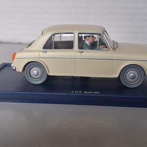 Op de afbeelding: Een beige oldtimer automodel met een bestuurdersfiguur erin, geplaatst op een donkerblauwe basis. De auto heeft grijze wielen en oranje achterlichten. De tekst "L'ILE NOIRE" is zichtbaar.