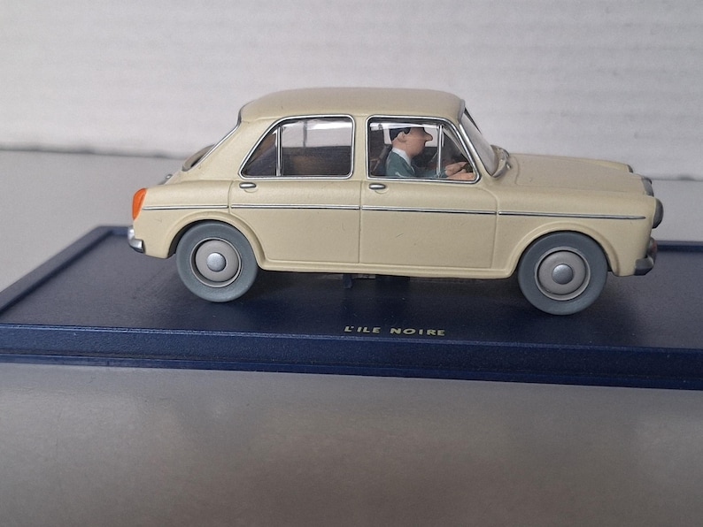 Op de afbeelding: Een beige oldtimer automodel met een bestuurdersfiguur erin, geplaatst op een donkerblauwe basis. De auto heeft grijze wielen en oranje achterlichten. De tekst "L'ILE NOIRE" is zichtbaar.