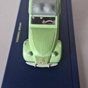 May include: A light green toy car with a gray top, displayed on a dark blue rectangular base. The car features a classic design with a ribbed front and the Citro&euml;n logo. The text "CAPITAINE TOURNESOL" is visible on the base.