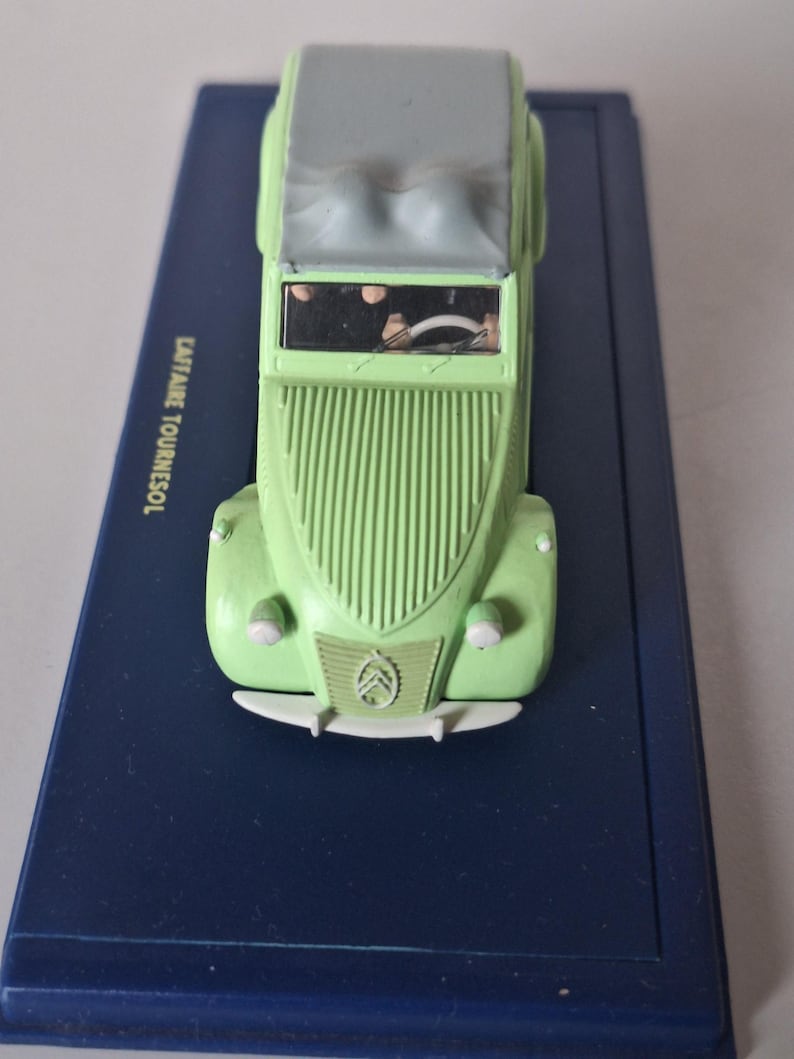 May include: A light green toy car with a gray top, displayed on a dark blue rectangular base. The car features a classic design with a ribbed front and the Citro&euml;n logo. The text "CAPITAINE TOURNESOL" is visible on the base.