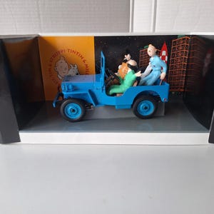 May include: A blue toy jeep with figures of characters from the Tintin comic series. The jeep is in a display box with a backdrop of a city and space. The figures include Tintin, Captain Haddock, and others.