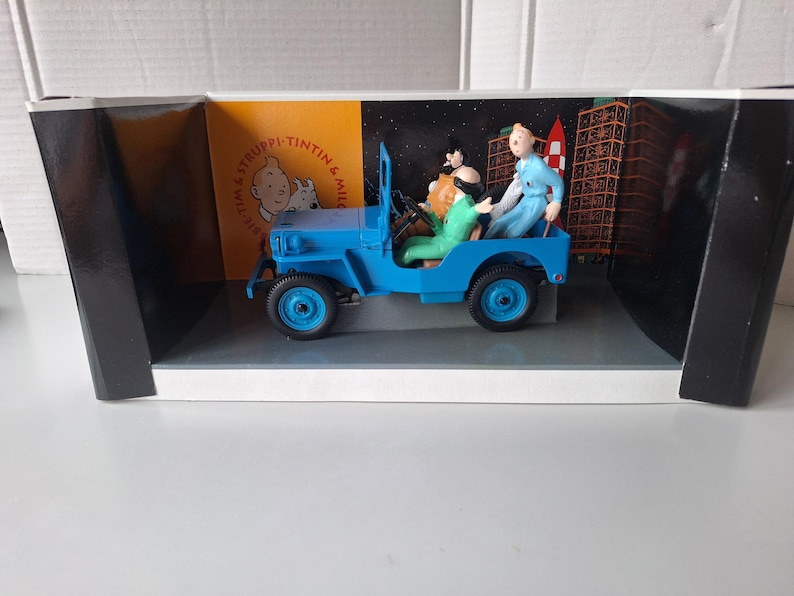 May include: A blue toy jeep with figures of characters from the Tintin comic series. The jeep is in a display box with a backdrop of a city and space. The figures include Tintin, Captain Haddock, and others.