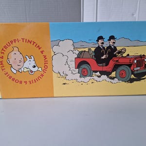 May include: A rectangular box featuring illustrations from the Tintin comic series. The left side has a portrait of Tintin and Snowy, with text in a circle. The right side shows two men in a red jeep driving through a desert landscape.
