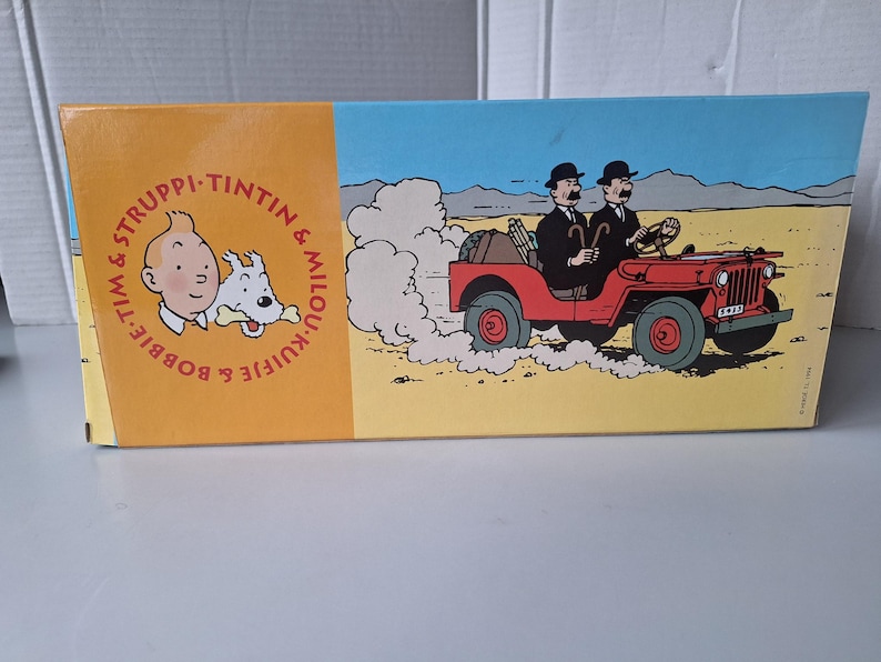 May include: A rectangular box featuring illustrations from the Tintin comic series. The left side has a portrait of Tintin and Snowy, with text in a circle. The right side shows two men in a red jeep driving through a desert landscape.