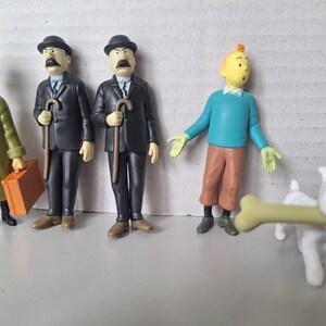 May include: A collection of Tintin figurines, including Tintin himself in a blue sweater and brown pants, and Snowy with a bone. Also featured are Thompson and Thomson, and a character in a green coat.