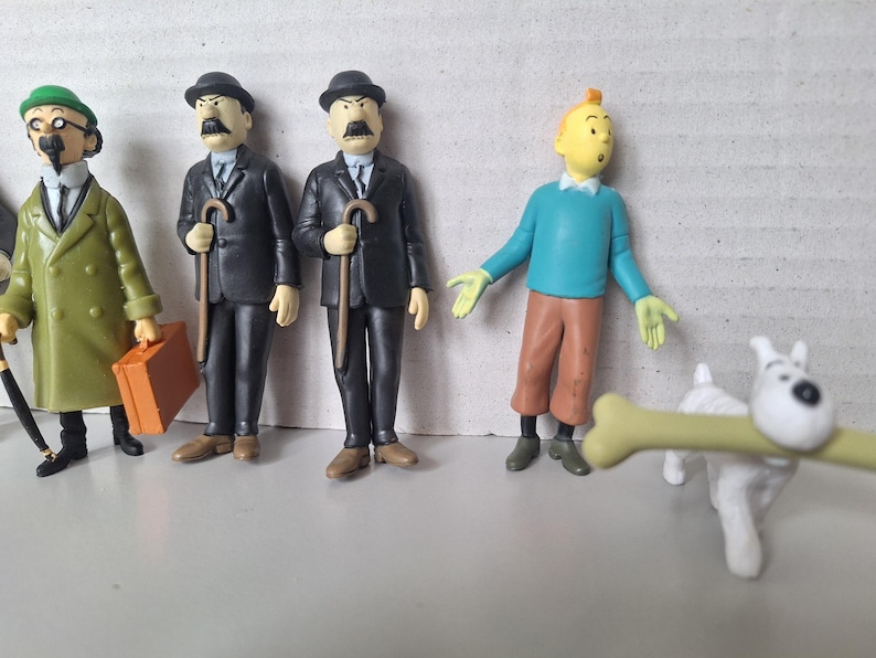 May include: A collection of Tintin figurines, including Tintin himself in a blue sweater and brown pants, and Snowy with a bone. Also featured are Thompson and Thomson, and a character in a green coat.