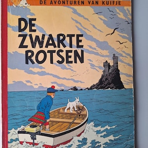 Op de afbeelding: Vintage stripboekcover getiteld "DE ZWARTE ROTSEN" met een jongen in een boot met een hond, varend naar een kasteel op een rotsachtig eiland. De cover heeft een rode rug en een blauwe en oranje lucht.