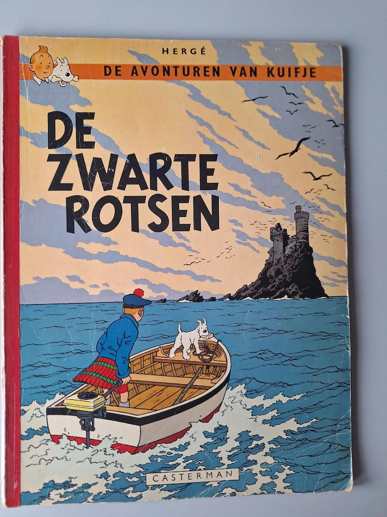 Op de afbeelding: Vintage stripboekcover getiteld "DE ZWARTE ROTSEN" met een jongen in een boot met een hond, varend naar een kasteel op een rotsachtig eiland. De cover heeft een rode rug en een blauwe en oranje lucht.