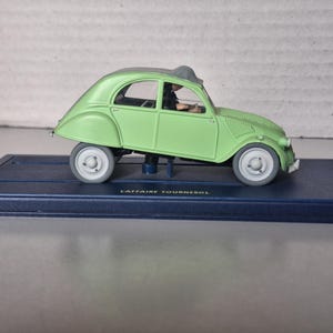 May include: A light green toy car model with white wheels and a dark gray roof, mounted on a dark blue rectangular base. The car has a classic design and is labeled "L'Affaire Tournesol".