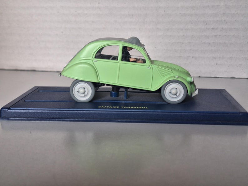 May include: A light green toy car model with white wheels and a dark gray roof, mounted on a dark blue rectangular base. The car has a classic design and is labeled "L'Affaire Tournesol".