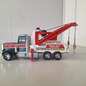 May include: A red and white toy tow truck with a green cab and a working crane. The truck has a white sign that reads "Heavy Duty Recovery Day & Night 52-7968".