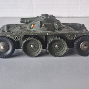 以下が含まれることがあります： 緑色の軍用装甲車の玩具。上部に大砲があり、4つの車輪がついています。車体の側面に小さな旗が取り付けられています。