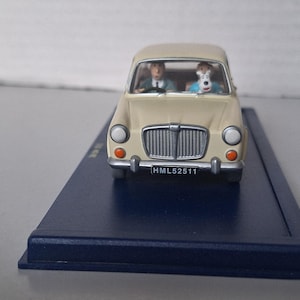 Op de afbeelding: Een cr&egrave;mekleurig model van een oldtimer met een bestuurder en een hond op de voorstoelen. De auto heeft een zwart kenteken met de tekst HML52511. Het model staat op een donkerblauwe basis.