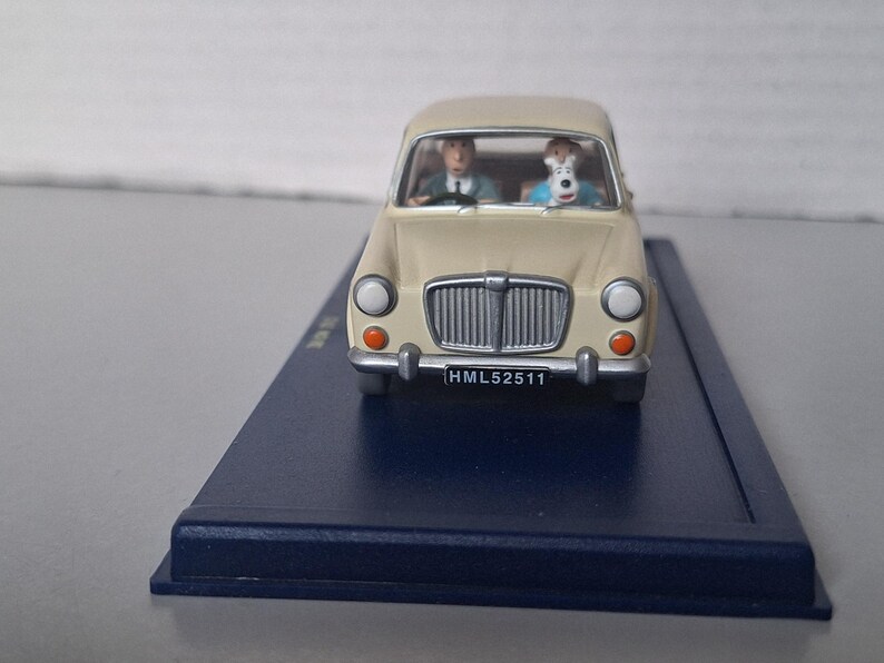 Op de afbeelding: Een cr&egrave;mekleurig model van een oldtimer met een bestuurder en een hond op de voorstoelen. De auto heeft een zwart kenteken met de tekst HML52511. Het model staat op een donkerblauwe basis.