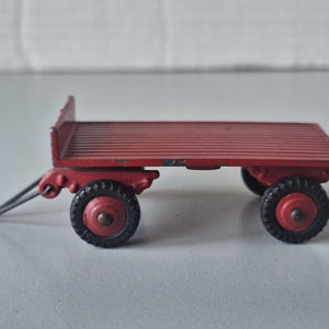 Può includere: Un rimorchio a pianale rosso giocattolo con un telaio in metallo e quattro ruote nere. Il rimorchio ha un gancio di traino in metallo e una barra di metallo che si estende dalla parte anteriore del rimorchio.