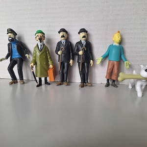 May include: A collection of Tintin figurines, including Captain Haddock, Thomson and Thompson, Tintin, and Snowy. The figures are brightly colored and stand on a gray surface. Snowy is holding a large bone.