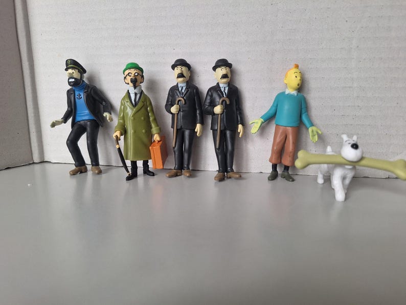 May include: A collection of Tintin figurines, including Captain Haddock, Thomson and Thompson, Tintin, and Snowy. The figures are brightly colored and stand on a gray surface. Snowy is holding a large bone.