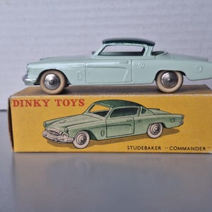 May include: A light green Dinky Toys Studebaker Commander die-cast model car with a dark green roof and white tyres, resting on its original yellow cardboard box featuring a green illustration of the car and the text DINKY TOYS STUDEBAKER COMMANDER.