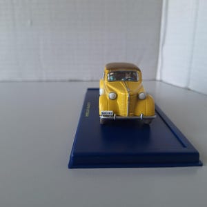 May include: A yellow vintage car model with a brown roof sits on a dark blue rectangular base. The car has a detailed front grille, headlights, and a license plate that reads "505191". The model is a collectible item.