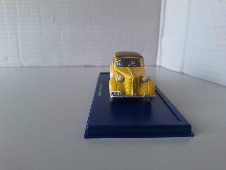 May include: A yellow vintage car model with a brown roof sits on a dark blue rectangular base. The car has a detailed front grille, headlights, and a license plate that reads "505191". The model is a collectible item.