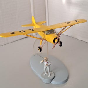May include: A yellow model aeroplane with black details and the letters "A D E" and "K H" on the wings. A small figurine of a man in a white suit stands below the plane on a grey base. The plane has a propeller and black wheels.