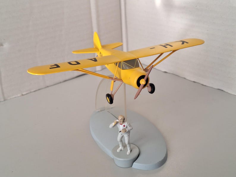 May include: A yellow model aeroplane with black details and the letters "A D E" and "K H" on the wings. A small figurine of a man in a white suit stands below the plane on a grey base. The plane has a propeller and black wheels.