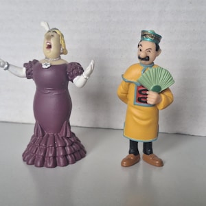May include: Two figurines: a woman in a purple dress with arms outstretched and a man in a yellow outfit holding a green fan. The woman has blonde hair and is singing. The man has a mustache and is wearing a hat.