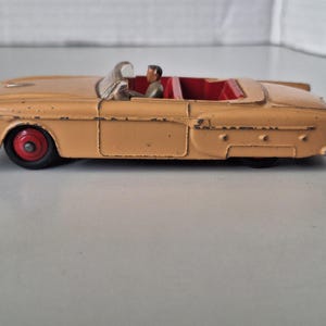 Op de afbeelding: Een vintage speelgoed cabriolet in lichtbruine kleur met rode binnenkant en wielen. De speelgoedauto heeft twee kleine figuren binnenin. De auto heeft een klassiek ontwerp.