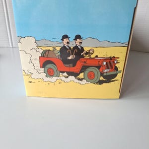 May include: A box featuring a cartoon illustration of two men in black suits and bowler hats driving a red jeep through a desert landscape. The jeep kicks up dust, and the sky is blue.