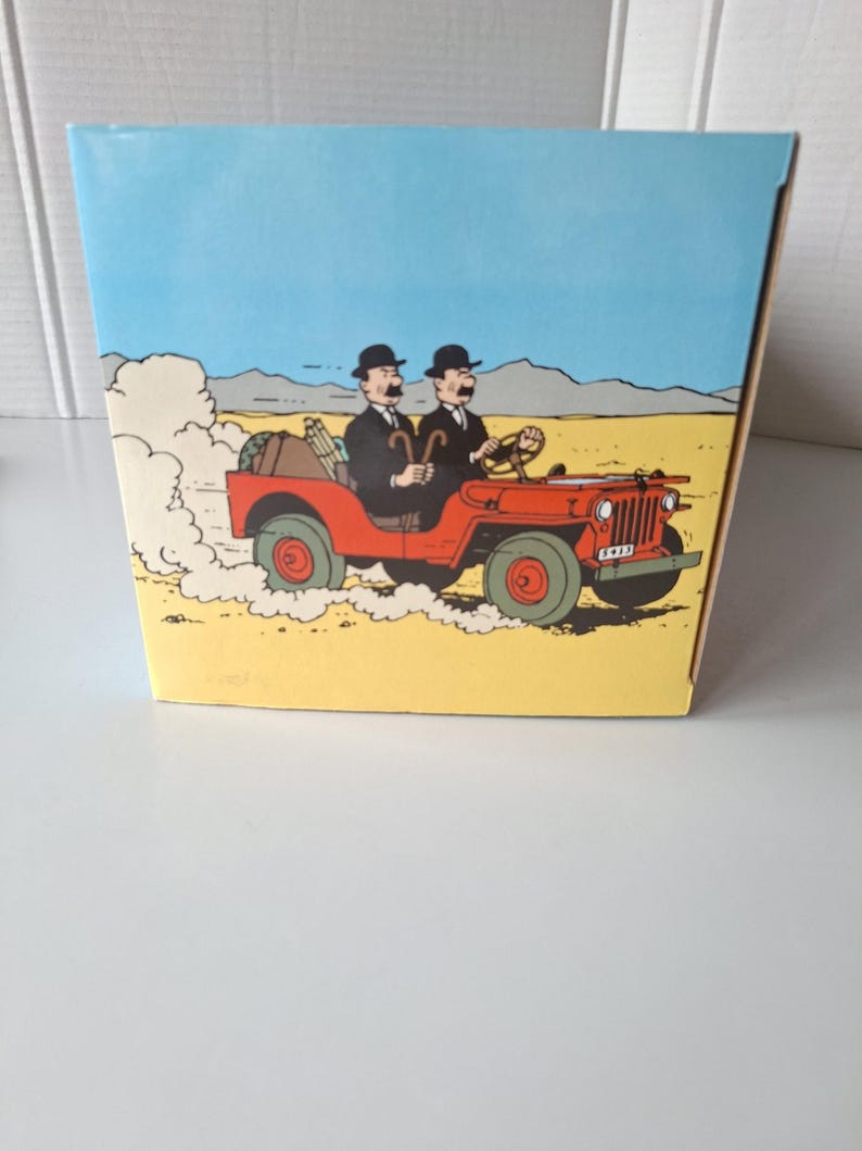 May include: A box featuring a cartoon illustration of two men in black suits and bowler hats driving a red jeep through a desert landscape. The jeep kicks up dust, and the sky is blue.