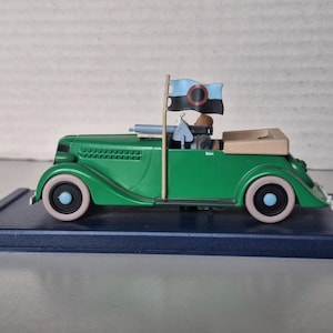 May include: A green vintage convertible toy car with cream-coloured tyres and a tan interior. The car has a flag on top with a black and blue design. The car is on a dark blue base.