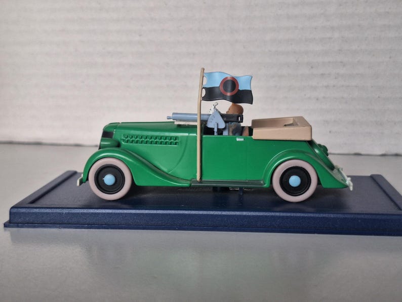 May include: A green vintage convertible toy car with cream-coloured tyres and a tan interior. The car has a flag on top with a black and blue design. The car is on a dark blue base.