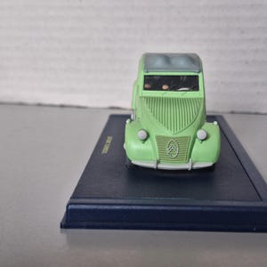 May include: A miniature green Citro&euml;n car model with a gray roof, displayed on a dark blue rectangular base. The car has a distinctive front grille and headlights. The model is a collectible item.