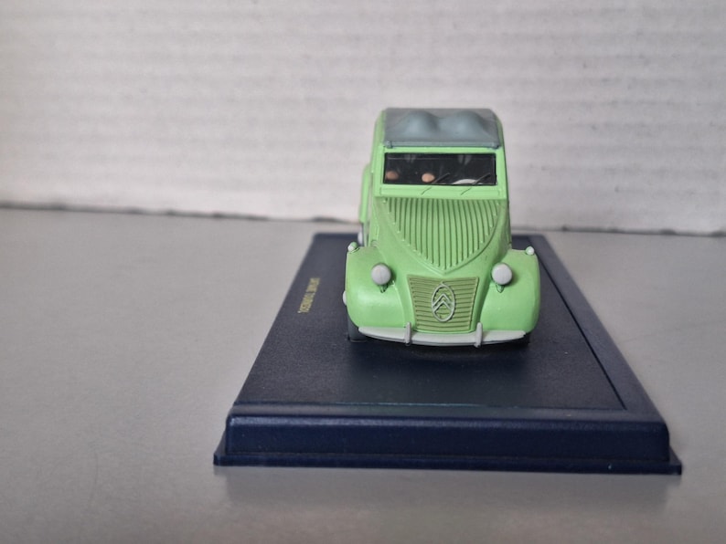 May include: A miniature green Citro&euml;n car model with a gray roof, displayed on a dark blue rectangular base. The car has a distinctive front grille and headlights. The model is a collectible item.