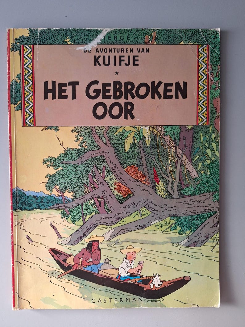 May include: A vintage comic book cover with the title "Het Gebroken Oor". The illustration shows a river scene with two people in a boat, and a dog. The book is by Herg&eacute;. The cover has a beige and brown color scheme.