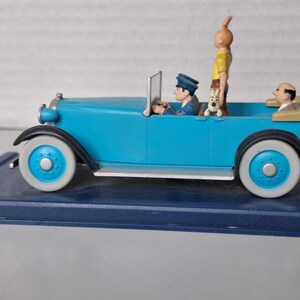 Puede incluir: Figura de coche antiguo azul claro con ruedas con borde blanco. El coche tiene un conductor, un pasajero y un perro. Una figura con pelo naranja est&aacute; de pie en la parte trasera. El coche est&aacute; sobre una base azul oscuro.