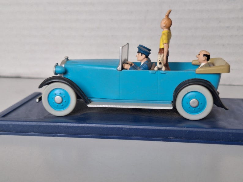 Puede incluir: Figura de coche antiguo azul claro con ruedas con borde blanco. El coche tiene un conductor, un pasajero y un perro. Una figura con pelo naranja est&aacute; de pie en la parte trasera. El coche est&aacute; sobre una base azul oscuro.