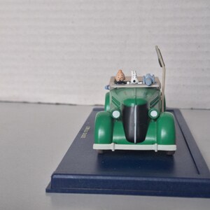 May include: A green vintage car model with a black front grill and white headlights. The car has a tan roof and features miniature figures of a man, a dog, and other items. The model sits on a blue rectangular base.