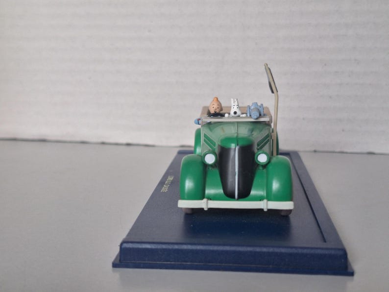 May include: A green vintage car model with a black front grill and white headlights. The car has a tan roof and features miniature figures of a man, a dog, and other items. The model sits on a blue rectangular base.