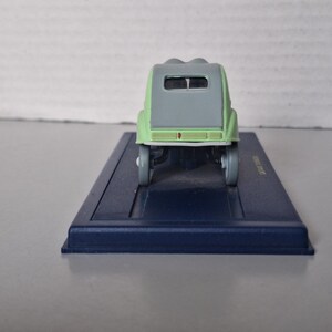May include: A miniature model of a light green and gray vintage car, viewed from the rear. The car sits on a dark blue rectangular base. The model has a rounded roof and small wheels.