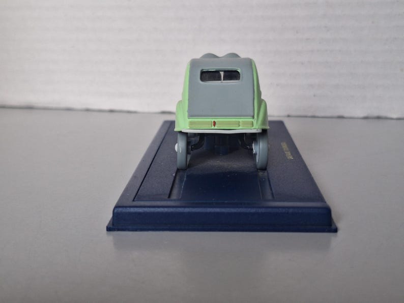 May include: A miniature model of a light green and gray vintage car, viewed from the rear. The car sits on a dark blue rectangular base. The model has a rounded roof and small wheels.