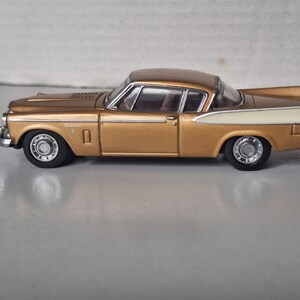 May include: A gold and cream-coloured die-cast model car featuring a two-tone paint scheme, chrome-coloured wheels, and a detailed exterior body with a long bonnet, a passenger cabin with visible windows, and rear tail fins in a classic coupe design.