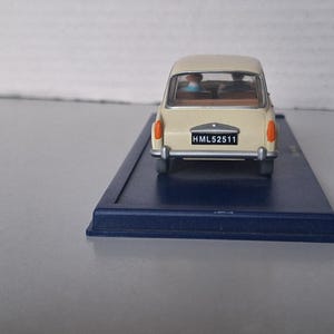 Op de afbeelding: Een cr&egrave;mekleurige miniatuurauto met oranje achterlichten en een zwart kenteken met de tekst "HML52511". De auto staat op een donkerblauwe rechthoekige basis. De ramen van de auto tonen twee figuren binnenin. Een klassiek automodel.
